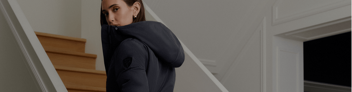 A woman wearing a dark navy puffer jacket stands on a wooden staircase inside a modern home, looking back over her shoulder.