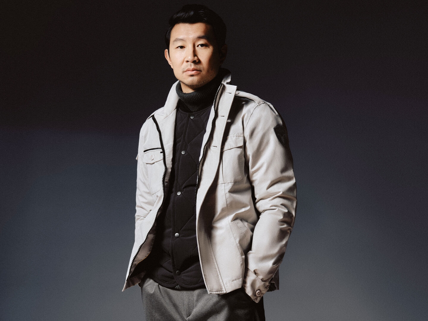 Simu Liu stands in a tonal grey environment and wears the Admiral Men's Shirt Jacket in the Light Grey colour. Underneath, Liu wears the Speck Men's Tailored Mid Layer Jacket in the Black colour.