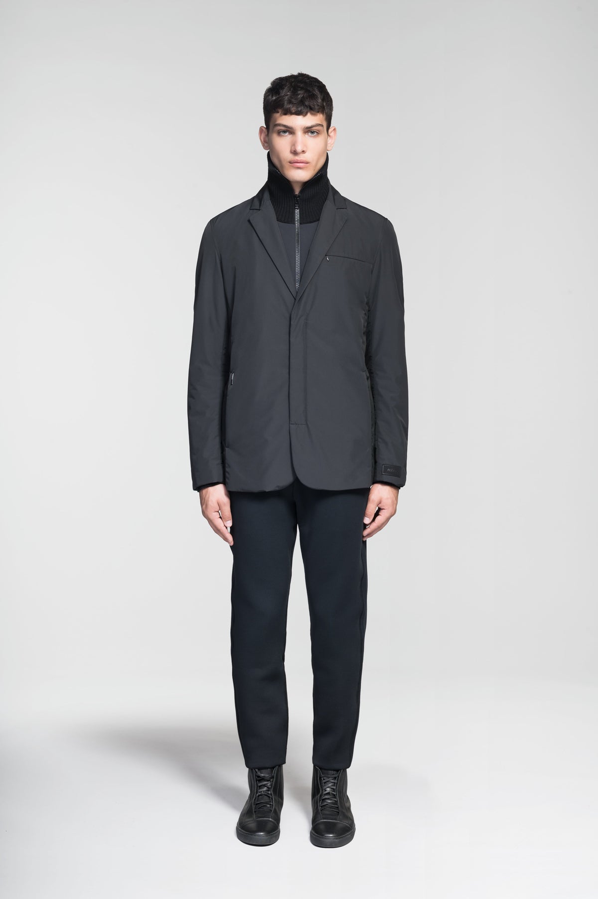 Cody Men's Tailored Travel Blazer in 3-ply micro denier and stretch nylon fabrication with DWR coating, Primaloft Gold Insulation Active+, hidden two-way zipper at centre front with snap closure placket, three invisible exterior zipper pockets, double back pleats, and hidden snap placket at cuffs, in Black