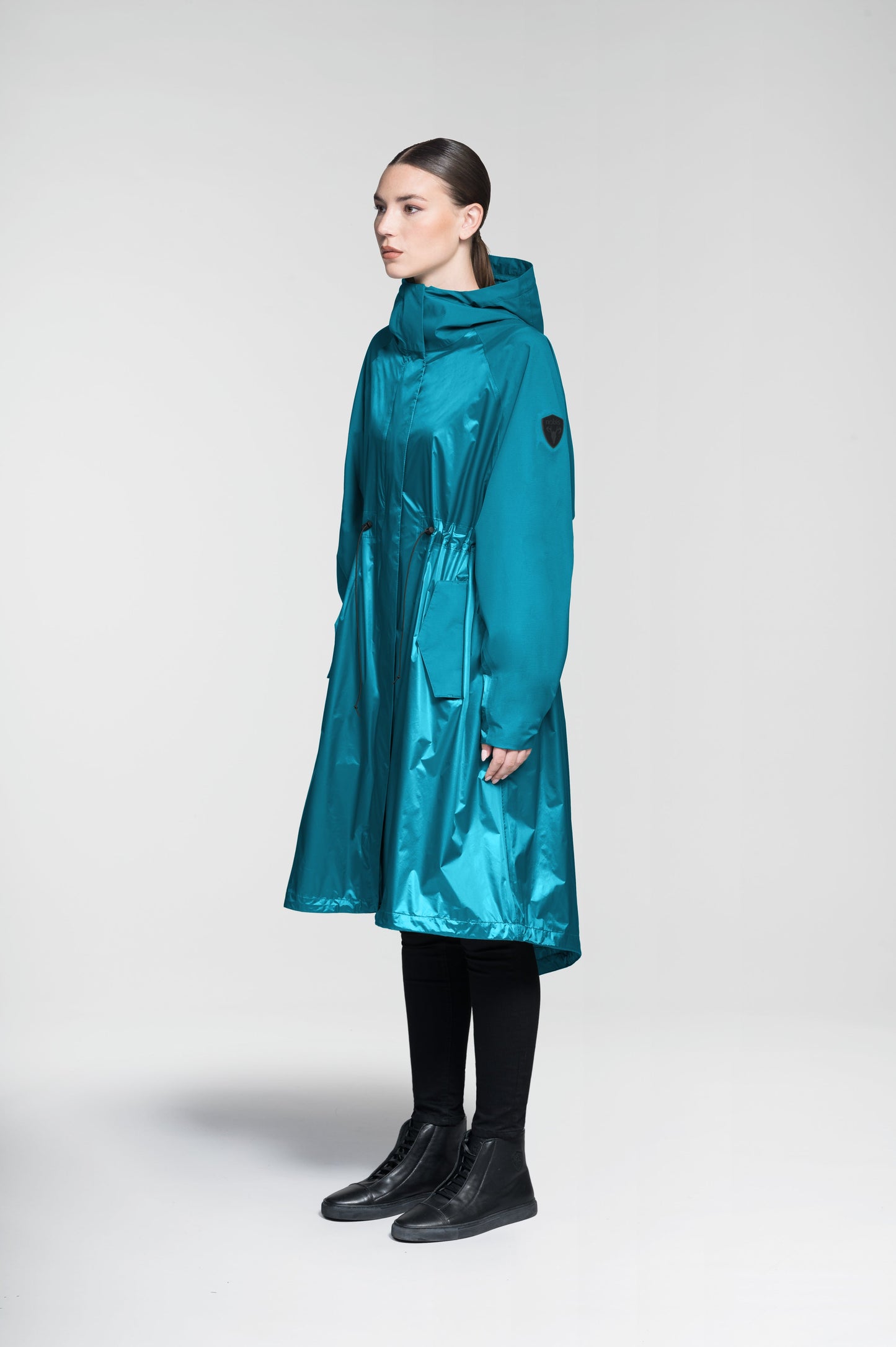 Reyna Women's Tailored Packable Long Shell Jacket in knee length, premium strech ripstop and contrast cire technical nylon taffeta fabrication, non-removable hood with adjustable draw cord, waist flap pockets with magnetic closures, adjustable draw cords at waist and bottom hem, adjustable snap cuffs, mesh back ventilation, hidden packable pocket, snap button wind flap, two-way branded zipper at centre front, adjustable snap button fishtail yoke at back, and large interior zipper pocket, in Deep Lake